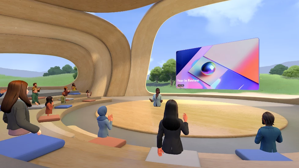 Avatar in white blazer standing at the center of a stage. Twelve other avatars seated in stadium seating facing the avatar in the center, who is standing in front of a "Year in Review" presentation.